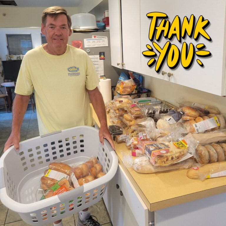 Photo of a volunteer from Food Link holding a basked of bread with a counter full of baked goods next to him